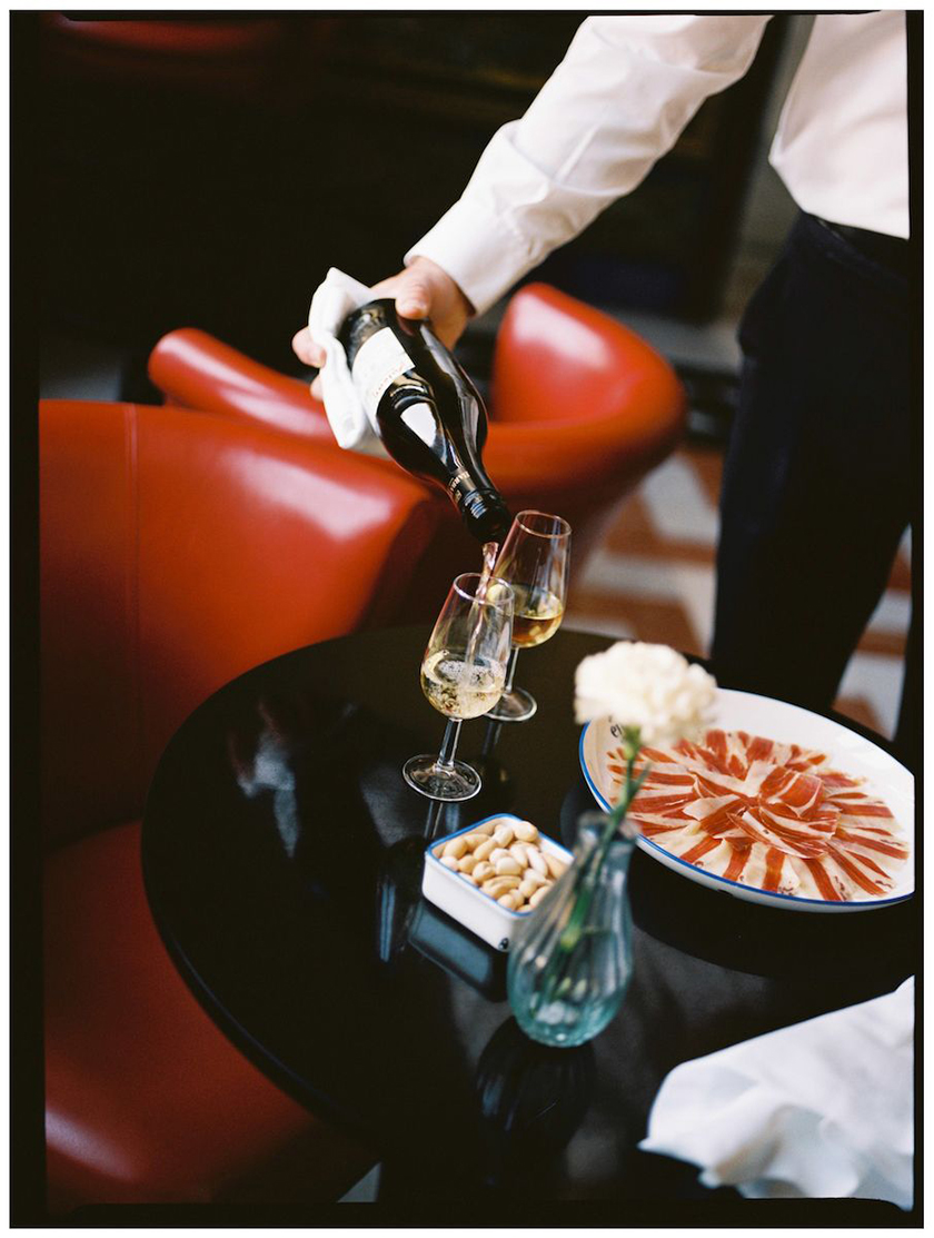 Waiter pouring a glass of wine from a table with a plate of ham and two glasses of wine.