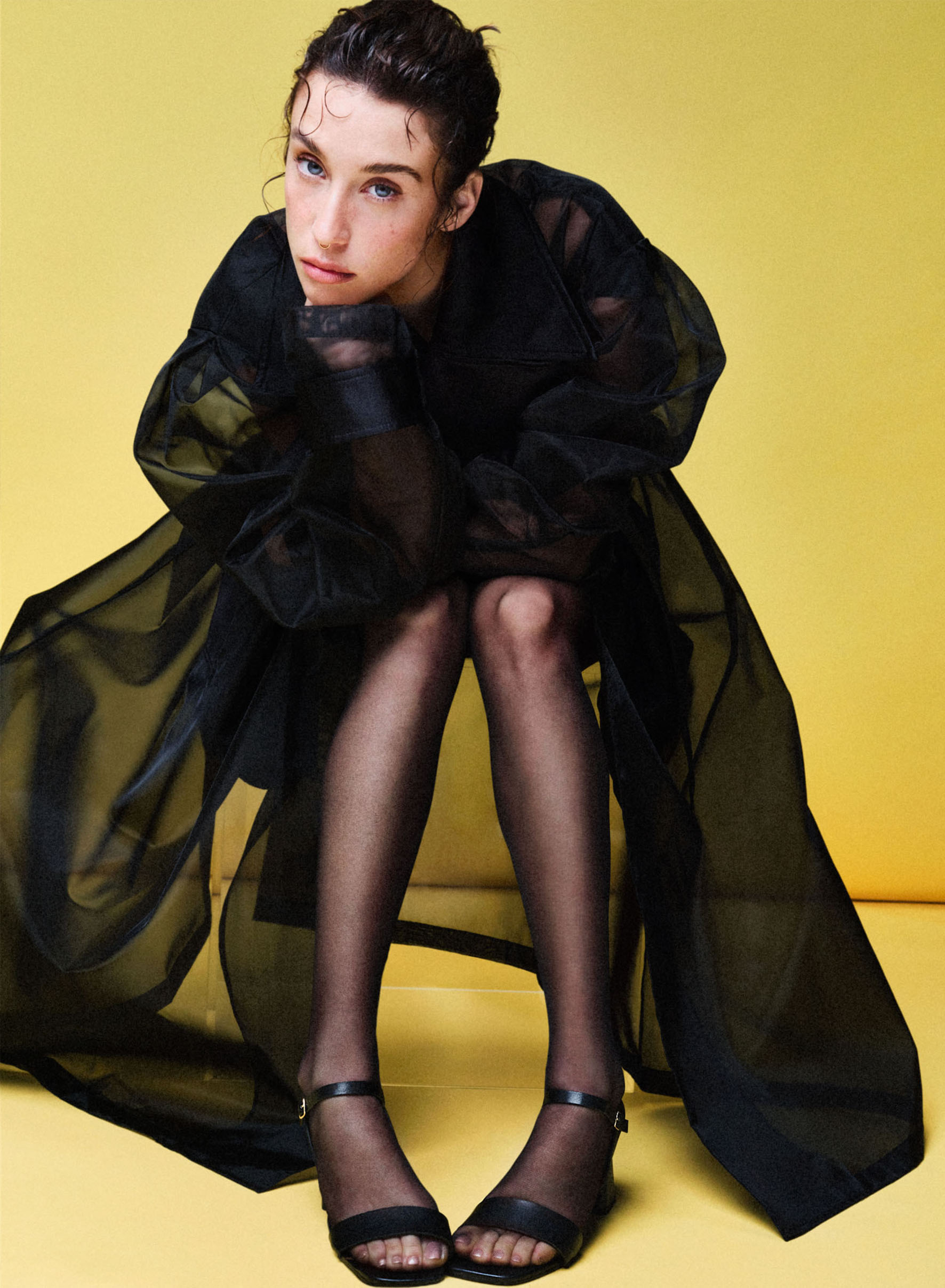 Maria Pedraza in black high heels sitting on a transparent object in a yellow background. 