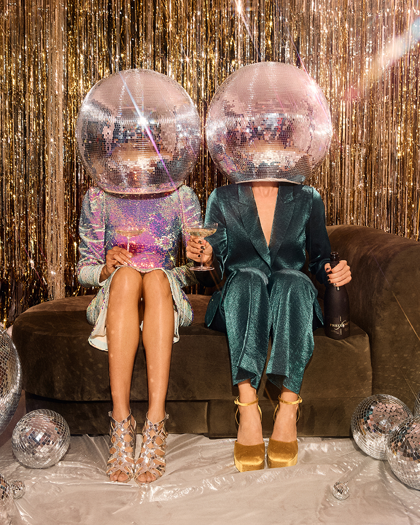 Two women sitting on a sofa, holding a bottle of freixenet - Cordón negro and a disco ball on their heads. 