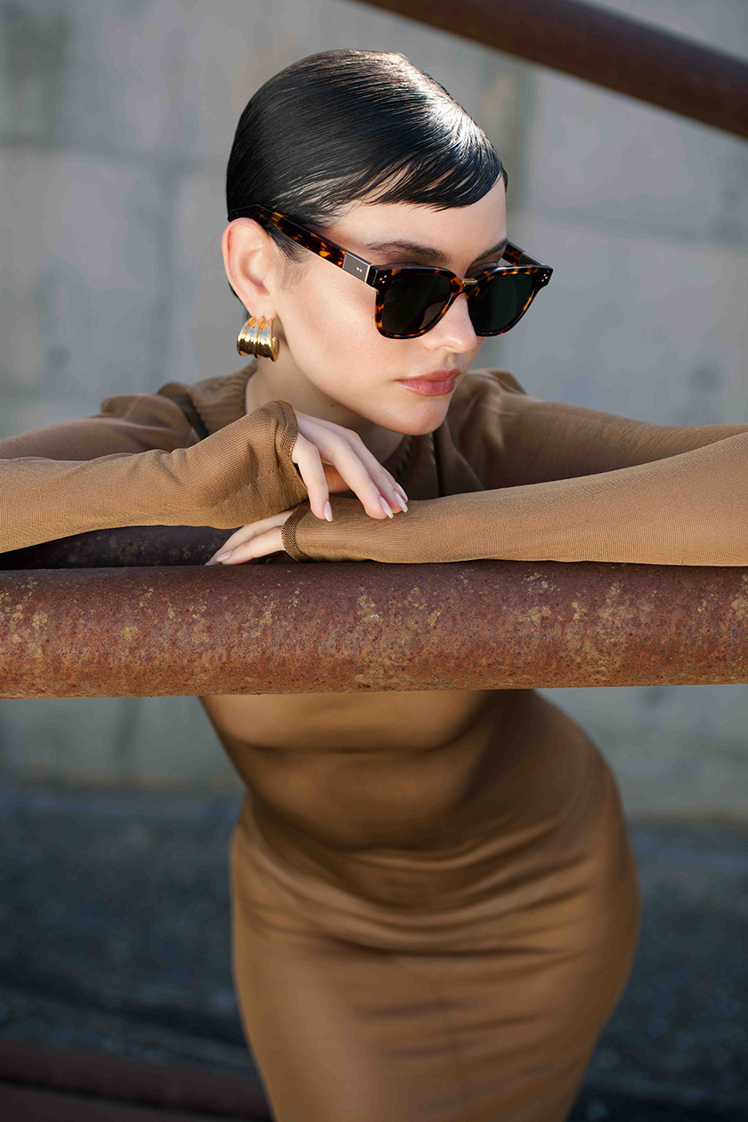 model leaning against a bar wearing a brown dress and glasses from the new Linda Farrow collection.