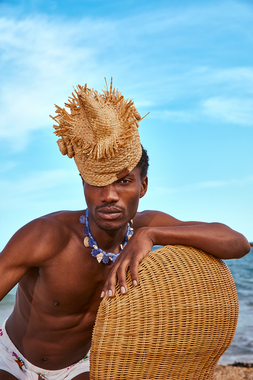 Model wearing a straw hat leaning against a chair with the sea in the background for the Pardo Hats campaign.
