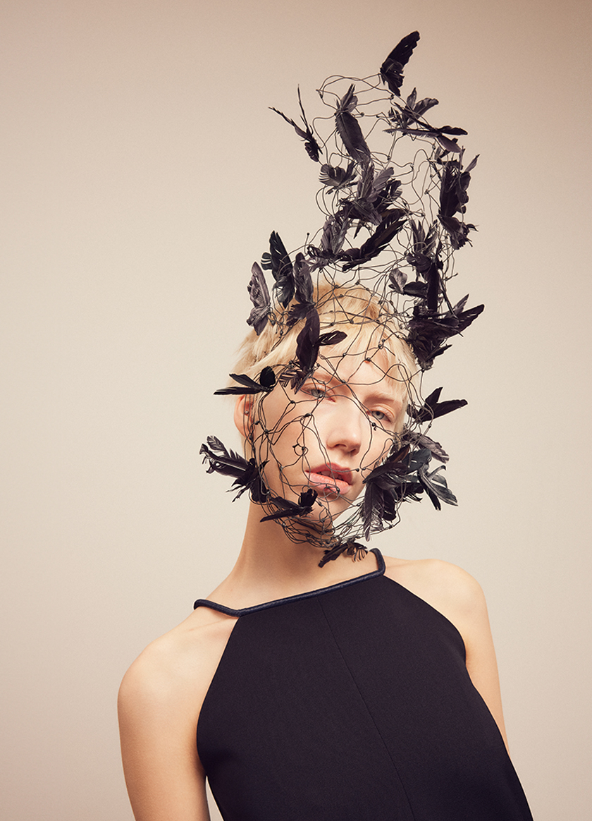 A woman in a dress with a bird cage on her head made of wires and feathers looks at the camera.