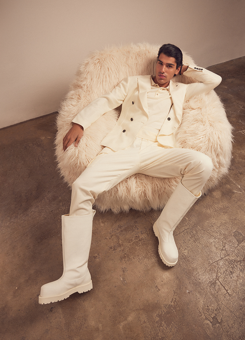 A man dressed in a white suit and white boots is stretched out on a white pouf looking at the camera.