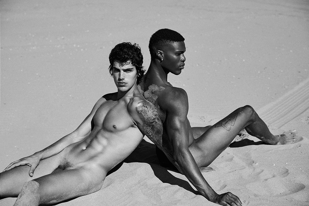 Two naked boys sitting on the sand of the beach sitting and leaning their backs. 