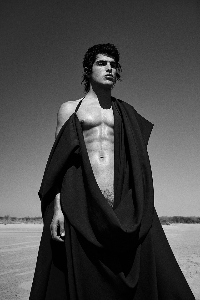 A shirtless boy in a black cape gazes at the horizon on the beach.