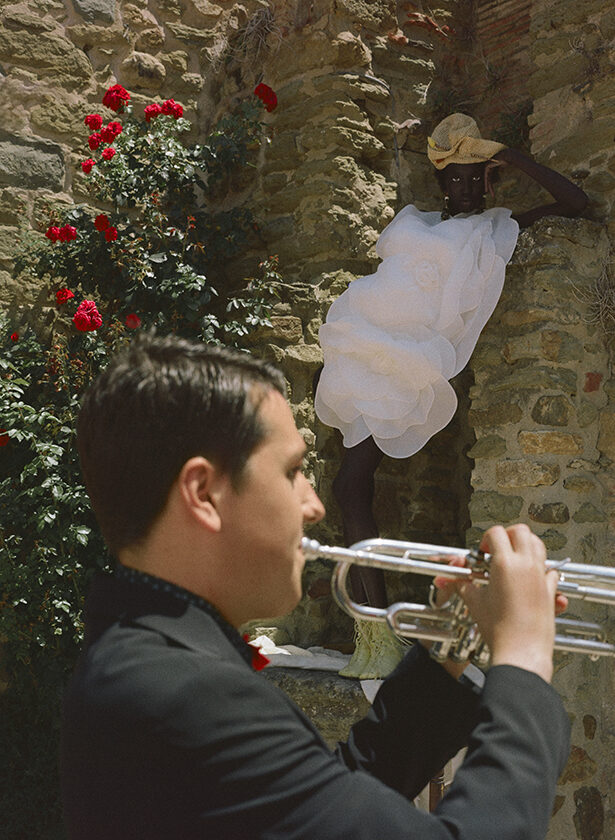 In the foreground a musician playing the trumpet and in the background the model Modleo Nyaueth leaning against a stone wall in a voluminous white dress for the magazine Numéro Netherlands. 