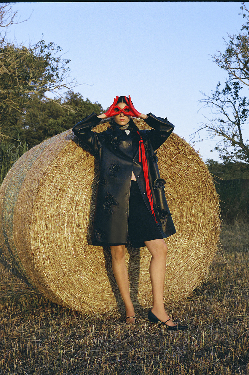 Model with a leather jacket and hands dipped in red ink resting on a bale of straw in the middle of a wheat field. 