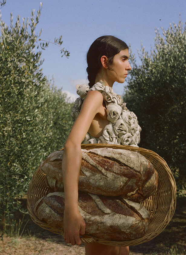 Model holding two large loaves of bread against a background of olive trees. 