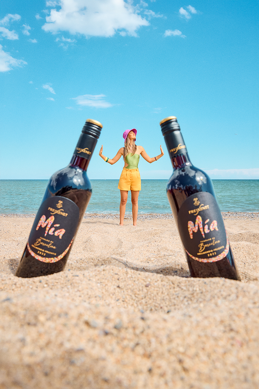 Two bottles of wine on the beach with a girl in the background playing with the size of the bottles.