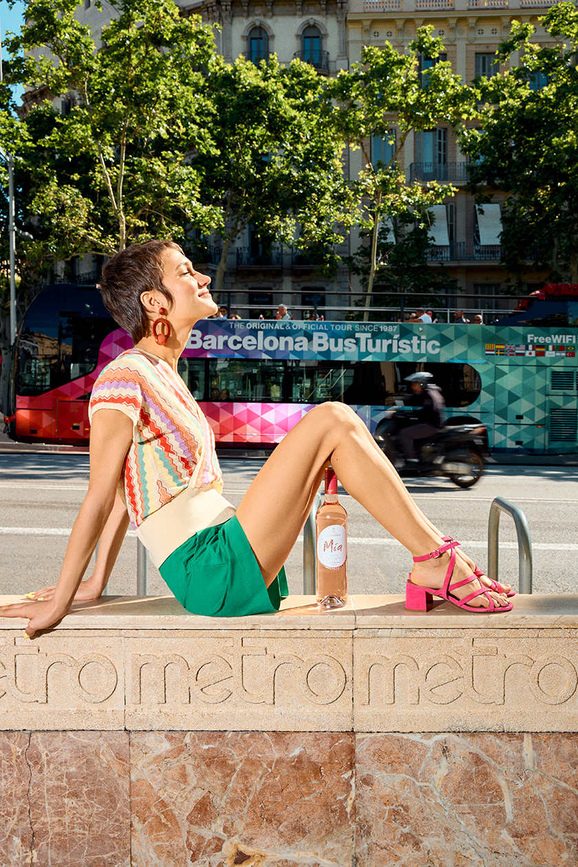 Seated model with a Barcelona bus in the background with a bottle of wine.